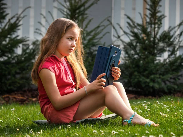 Distance learning during the quarantine period for the virus Covid-19. Cute little schoolgirl with long hair is studying from home, sitting in the garden on the grass. Uses a tablet and remote work via the Internet.