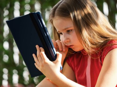 Covid-19 virüsünün karantina döneminde uzaktan öğrenme. Uzun saçlı sevimli küçük kız evden ders çalışıyor, bahçede çimlerin üzerinde oturuyor. İnternet üzerinden bir tablet ve uzaktan çalışma kullanır.