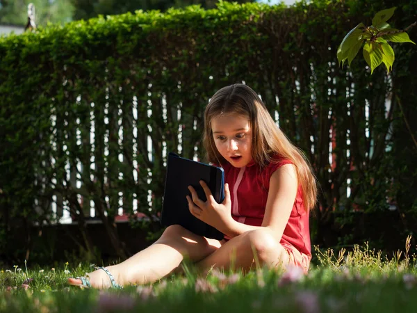 Distance learning during the quarantine period for the virus Covid-19. Cute little schoolgirl with long hair is studying from home, sitting in the garden on the grass. Uses a tablet and remote work via the Internet.