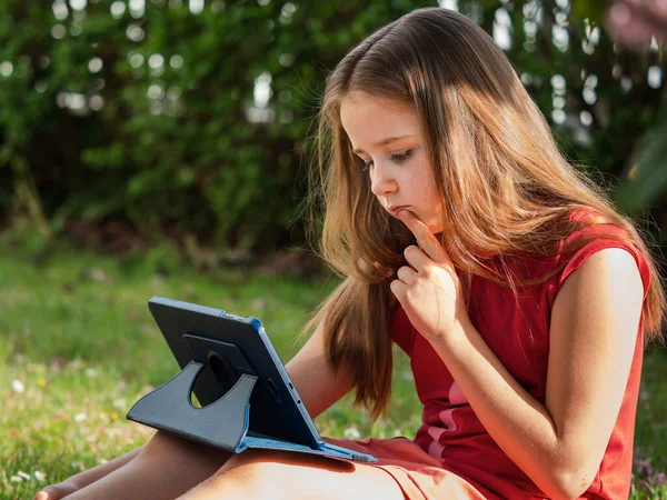 ウイルスCovid 19の隔離期間中の距離学習 長い髪をしたかわいい女の子は家から勉強していて 芝生の上の庭に座っています インターネットを介してタブレットやリモートワークを使用しています — ストック写真