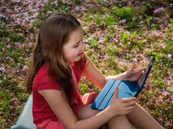 Distance learning during the quarantine period for the virus Covid-19. Cute little schoolgirl with long hair is studying from home, sitting in the garden on the grass. Uses a tablet and remote work via the Internet.