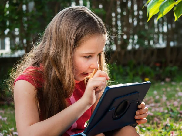 Distance learning during the quarantine period for the virus Covid-19. Cute little schoolgirl with long hair is studying from home, sitting in the garden on the grass. Uses a tablet and remote work via the Internet.