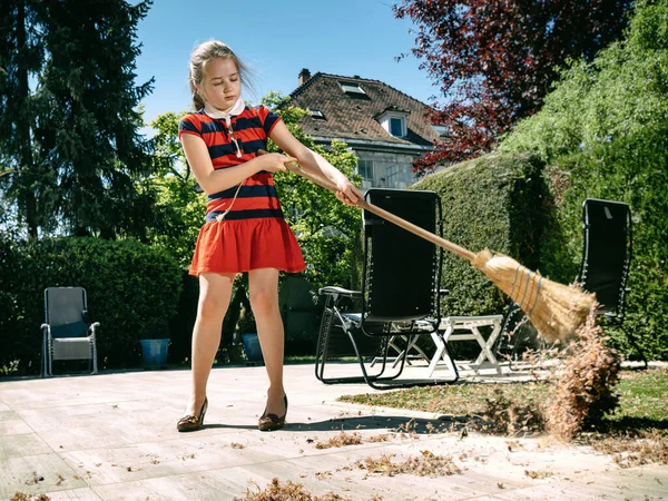 一个女学生在院子里摆姿势 手里拿着扫帚 穿着妈妈的鞋子 在花园里打扫卫生阳光灿烂的天气快乐的童年 — 图库照片