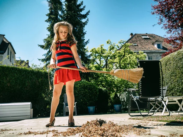一个女学生在院子里摆姿势 手里拿着扫帚 穿着妈妈的鞋子 在花园里打扫卫生阳光灿烂的天气快乐的童年 — 图库照片
