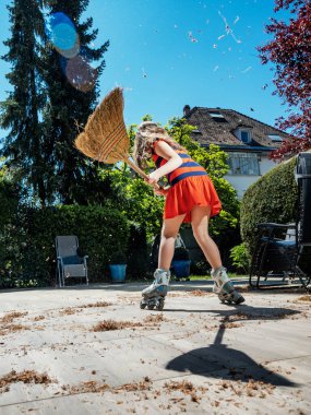 Bir kız öğrenci paten kayarken bahçede süpürgeyle poz veriyor. Hizmetçi. Bahçeyi süpürüyor. Temizliğin otomasyonu ve bunu eğlenceye dönüştürmek. Güneşli bir hava. Neşeli çocukluk.