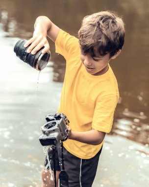 Okullu bir çocuk nehirde dikiliyor ve aynasız bir fotoğraf makinesini yok ediyor. Teknolojinin eskimesi kavramı. Fotoğrafçılar günü, mizah. Tonlu resim. Yumuşak ışık fotoğrafı.
