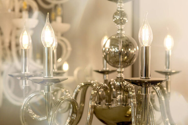 The picture of a glowing chandelier with metal chrome fixtures and shades in the form of candles. The concept of the classical style in the interior. Close up. Soft warm filter, selective focus.