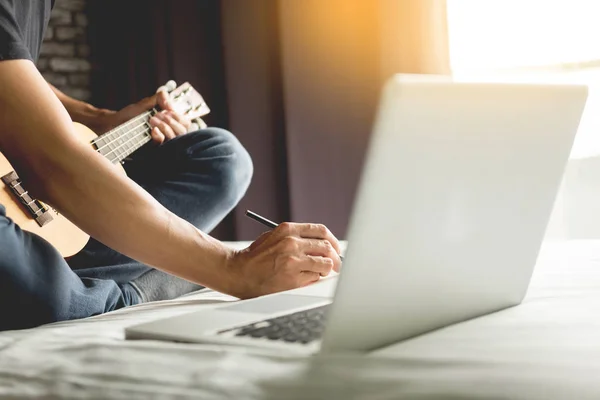 Glad ung asiatisk man spela ukulele sitter på sängen i sovrummet. — Stockfoto