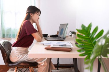 Çekici, genç, güzel Asyalı kadın ev içi oturma odasında bağımsız olarak otururken, uzaktan ya da ev kavramından bağımsız olarak çalışırken dizüstü bilgisayar ve belgeyle çalışıyor.