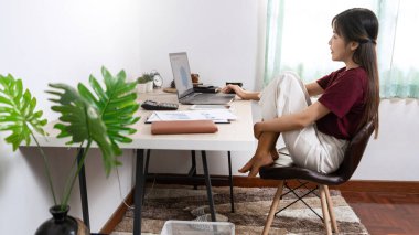 Çekici genç Asyalı kadın girişimci ya da evde dizüstü bilgisayarla çalışan serbest çalışan iş raporları ve oturma odasındaki kanepede internetten iletişim kuran, uzaktan erişim kavramına uzaktan erişen