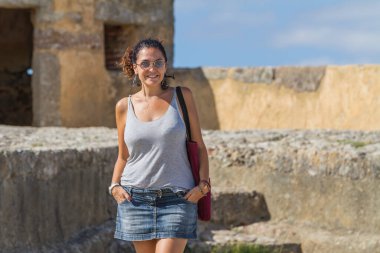 Mujer model en jeans cortos y camiseta gris sin mangas y gafas de sol, caminando entre las paredes de una fortaleza de piedra
