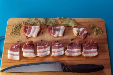 Yatay fotoğrafın mavi arka planında tahta bir kesim tahtası resmi var. Parçalara ayrılmış taze et, et için kurutulmuş bitkisel baharatlar, karabiber, körfez yaprağı.