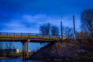 Yatay fotoğraf bir kentsel alanın resmini gösteriyor: ağaçlar, bir köprünün parçası ve iki fener. Sessiz bir nisan akşamı