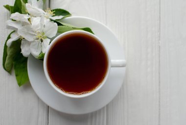 Beyaz porselen fincanda sıcak çay, beyaz çiçekli elma dalı beyaz ahşap arka plan.