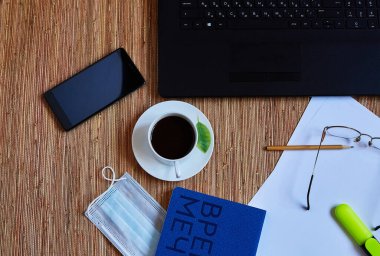 Bilgisayarlı çalışma alanı, koruyucu maske, mavi defter, kahve ve akıllı telefon 