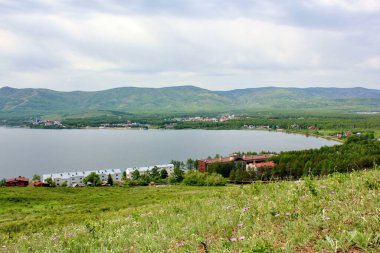 Harika bir panoramik manzara. Görkemli dağların ortasında sisli, temiz bir dağ gölü olan bir göl. Yeşil çimenler, sarı ve mavi çiçekler ön planda.