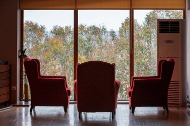 Sonbahar ağaçlarının manzaralı büyük pencerenin önünde üç kırmızı rahat koltuk ve klima.