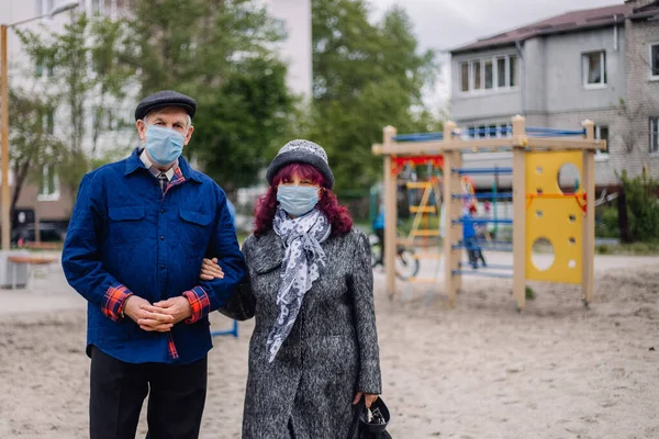 Elderly couple in protective medicine masks outdoors. Old people with covid protection on faces in town yard. Children playground background