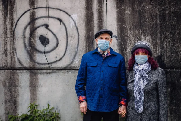 Elderly couple in protective medicine masks outdoors. Old people with covid protection on faces in town yard. Street background