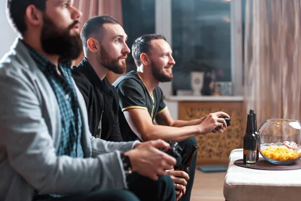 Uma companhia de homens se divertindo e rindo sentados no sofá em casa com cerveja e batatas fritas, dois deles com joysticks na mão jogando videogame de computador — Fotografia de Stock