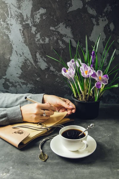 Dziewczyna ręce zbliżenie z piórem w ręku pisania w Notatniku. Na stole jest filiżanką kawy i wazon z kwiatami krokusy — Zdjęcie stockowe