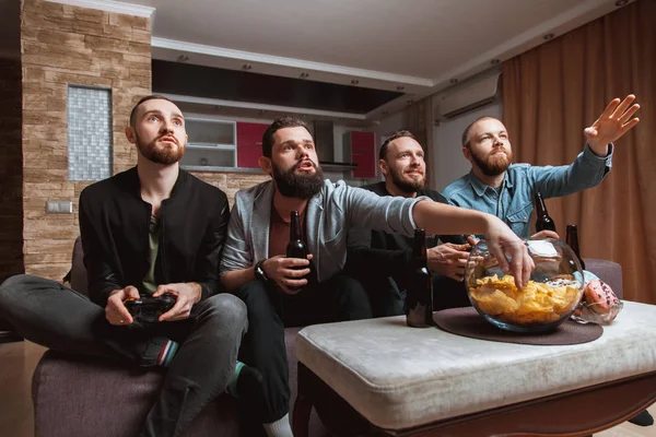 Männer mit Bart sitzen zu Hause auf der Couch, Bier und Steuerknüppel in der Hand, um Computervideospiele zu spielen — Stockfoto