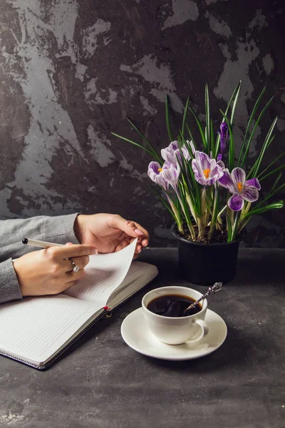 The girl's hands closeup with a pen in hand writing in a notebook. On the table is a Cup of coffee and a vase with flowers crocuses — Stock Photo, Image