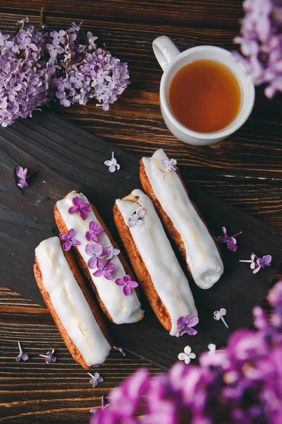 Красиві клерки з кремом і чашкою чаю на дерев'яній дошці з бузковими квітами — стокове фото