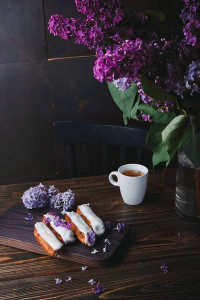 Krásný éclair dorty se smetanou a šálek čaje na dřevěné desce s fialovými květy — Stock fotografie
