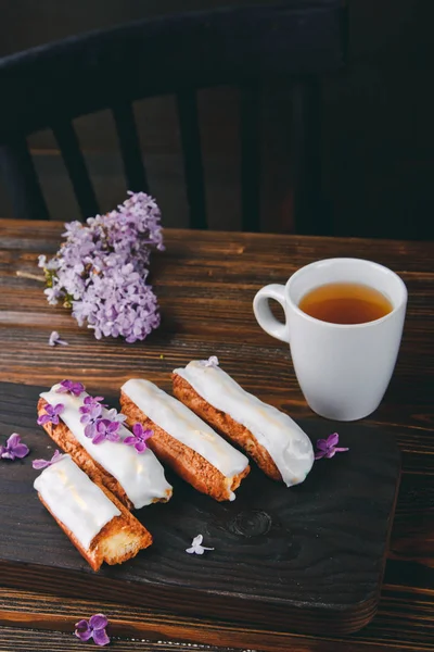 Красиві клерки з кремом і чашкою чаю на дерев'яній дошці з бузковими квітами — стокове фото