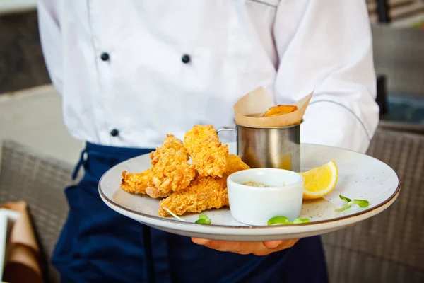 Peixe e batatas fritas em um prato branco nas mãos dos chefs do restaurante — Fotografia de Stock