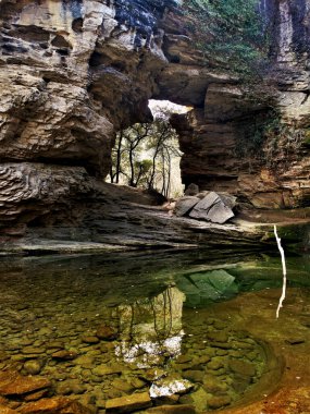 Manzara, büyük bir kayanın içindeki deliği ve Katalonya 'da La Forada denilen bir göldeki suyun yansımasını gösteriyor.