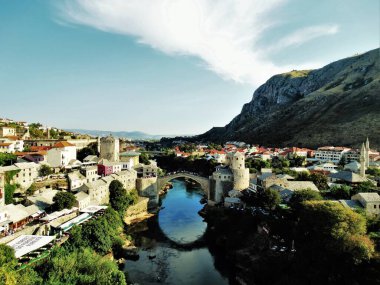 Bosna 'daki Mostar şehri, güneşli bir yaz gününde eski bir kasabayı, bir ortaçağ köprüsünü ve onun nehre yansımasını gösteriyor.