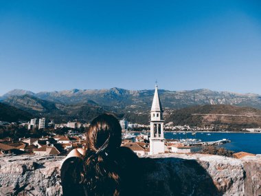 Karadağ 'da Budva' nın güzel sahil kasabasına bakan bir kız. Önünde bir kilise kulesi, önünde dağlar ve deniz var.