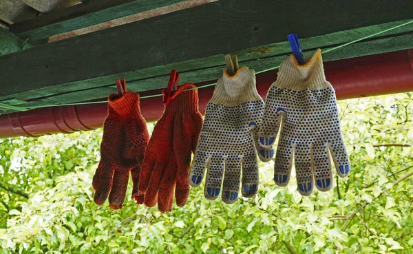 White and red work gloves are hung on a rope for drying under the roof against a background of green leaves on a bright day.