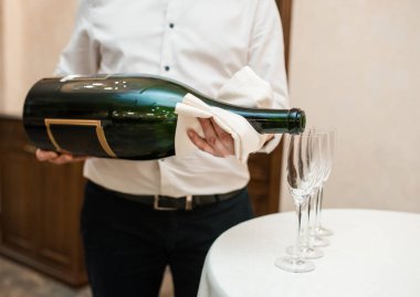 Garson büyük bir şişe şampanyayı şeffaf bir bardakta beyaz bir masanın üzerine koyuyor.