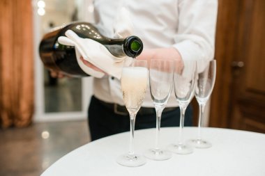 Garson büyük bir şişe şampanyayı şeffaf bir bardakta beyaz bir masanın üzerine koyuyor.
