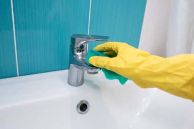 Metalik musluk, sarı lateks eldiven giyen biri tarafından yeşil süngerle temizleniyor.