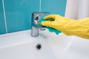 Metalik musluk, sarı lateks eldiven giyen biri tarafından yeşil süngerle temizleniyor.