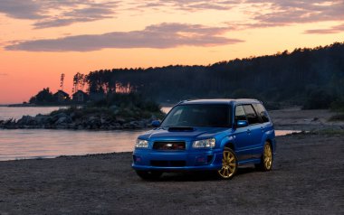 Mavi spor araba Subaru Forester STi Obskoe 'nun kıyısındaki su deposunda yaz günbatımında