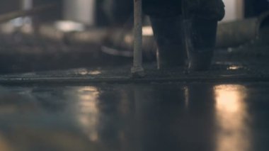 Çizmeli işçiler paspas ve kürekle beton kütleden pürüzsüz bir yüzey oluştururlar (işçinin yakın görüntüsü güneş ışığı bantlarındaki sıvı beton karışımından bir zemin oluşturur.)