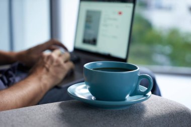 Adam elinde mavi bir fincan kahveyle dizüstü bilgisayarda çalışıyor.