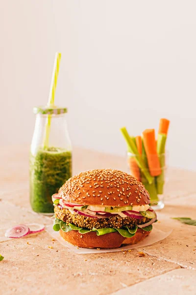 Bir şişe yeşil smoothie ile vejetaryen burger ve bardakta kerevizli havuç. Taş bej masa