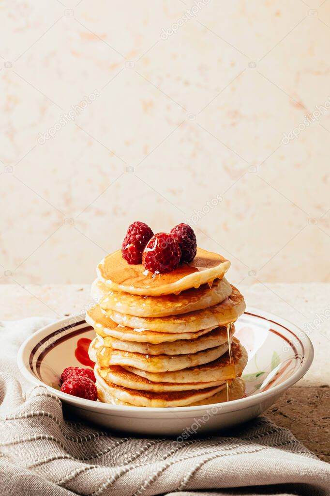 Pancakes poured with honey decorated with raspberries on a plate on a beige background. Stone table and textiles. Copy space