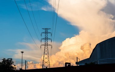 Yüksek voltajlı elektrik kablosu ve arka planda günbatımı gökyüzü olan fabrikanın silüeti..