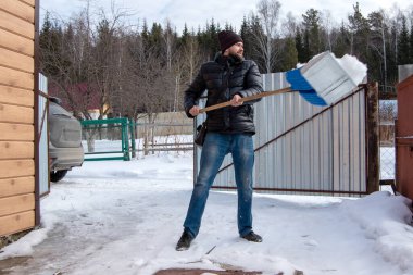 Siyah ceketli, kot pantolonlu ve siyah şapkalı beyaz bir adam park yerinde kar temizliyor..