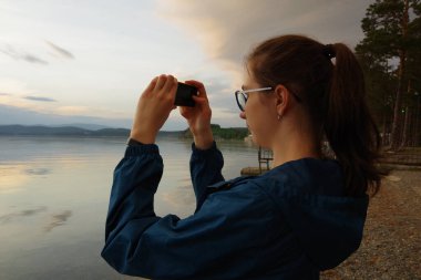 Mavi ceketli güzel kız elinde bir kamera tutuyor, gün batımında gölde ya da denizde fotoğraf çekiyor..
