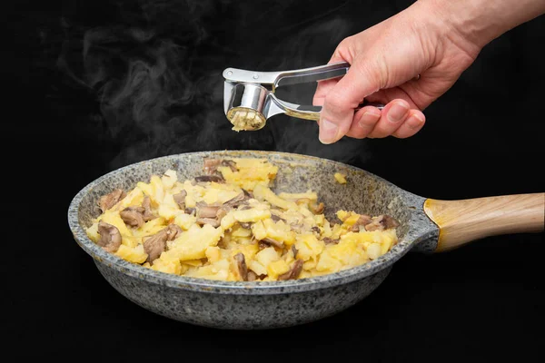 Female hand crushes garlic through a juicer in a fried potato with mushrooms in a gray with spots pan on a black table, close-up, black background