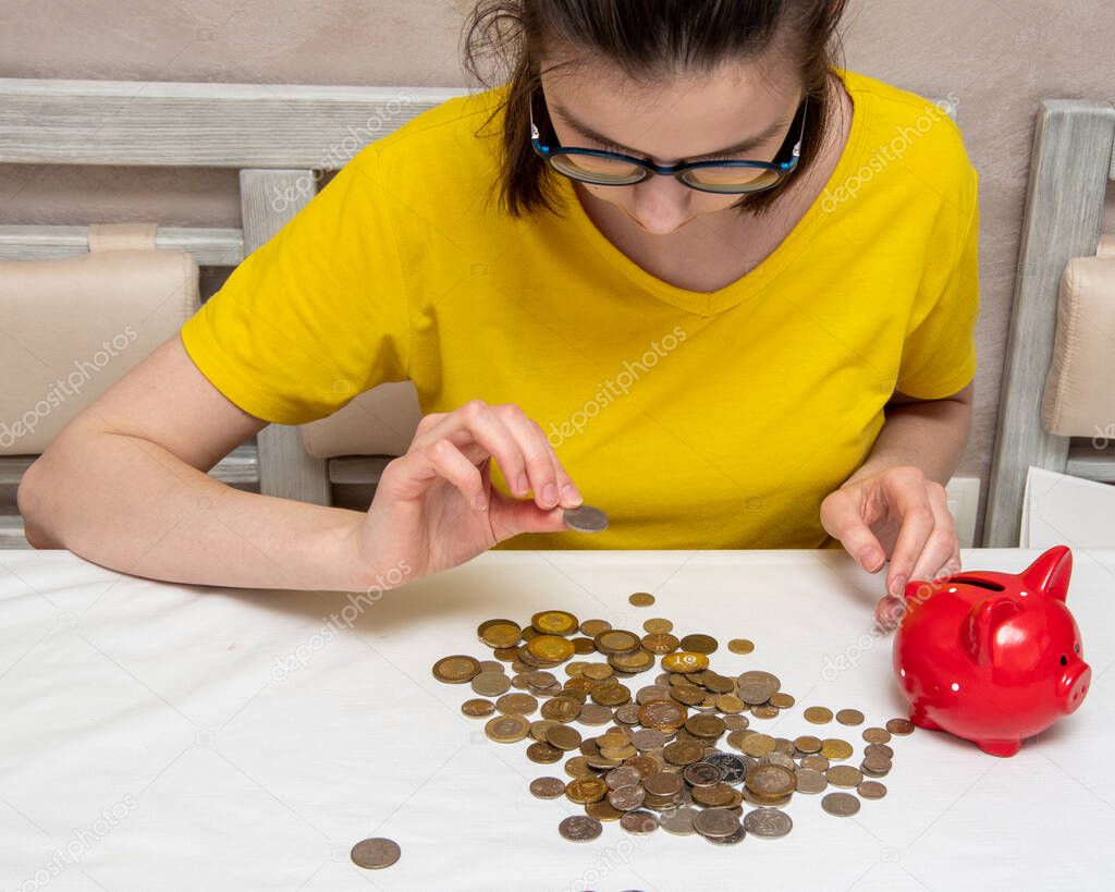 Portrait of a young girl in a yellow T-shirt looks at a coin, there are a lot of coins on the table, a red piggy bank.