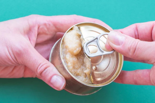 The hand of an adult man holds a can of canned food, the second hand opens the lid jerking a key. In the can of fish caviar. Top view on green background with copy space. Close-up